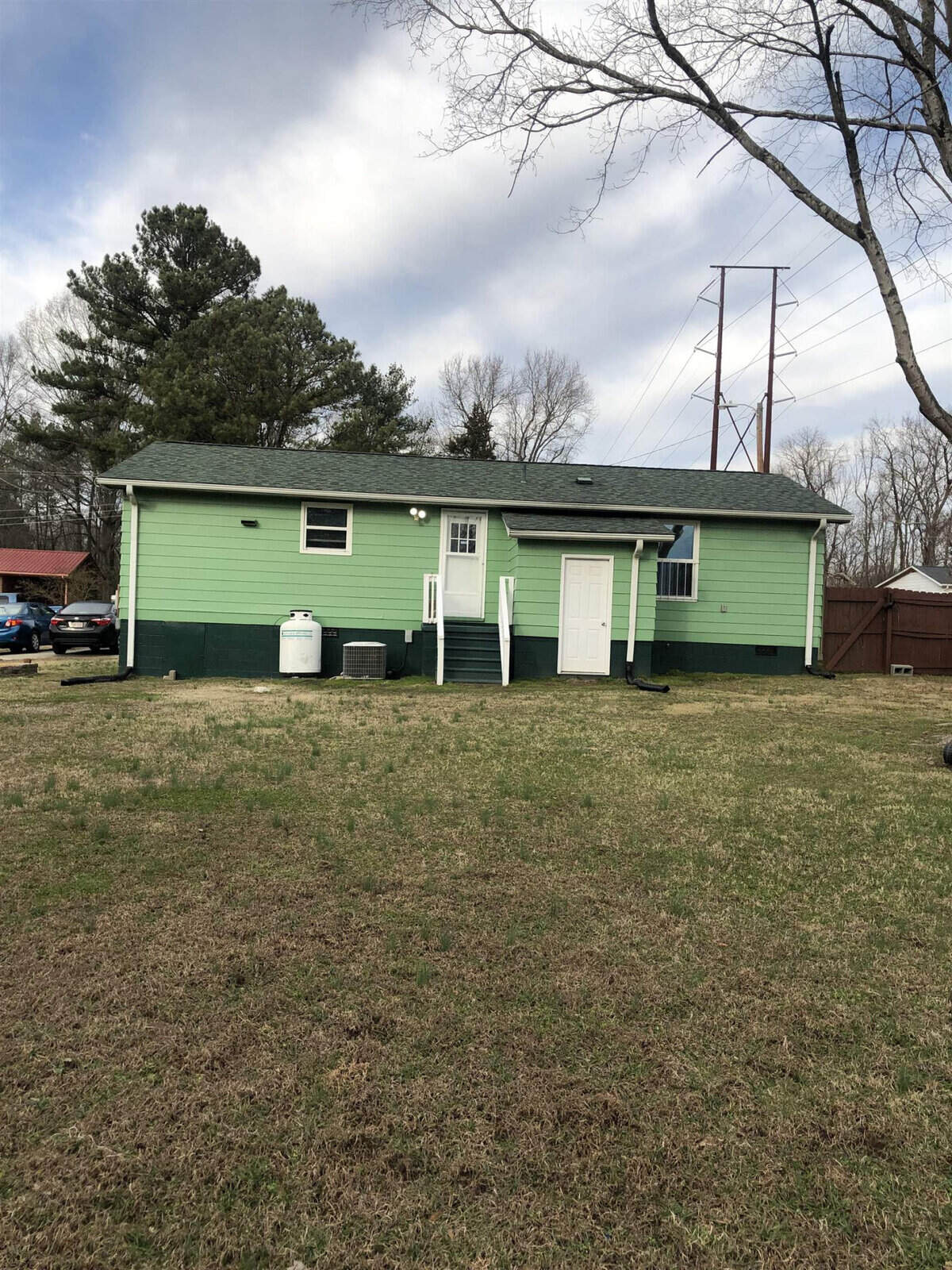 A single-story, green house with a white door and trim sits on a grassy yard. It has a small set of steps leading to the back door and a propane tank near the rear of the house. Trees and utility poles are visible in the background under a partly cloudy sky.