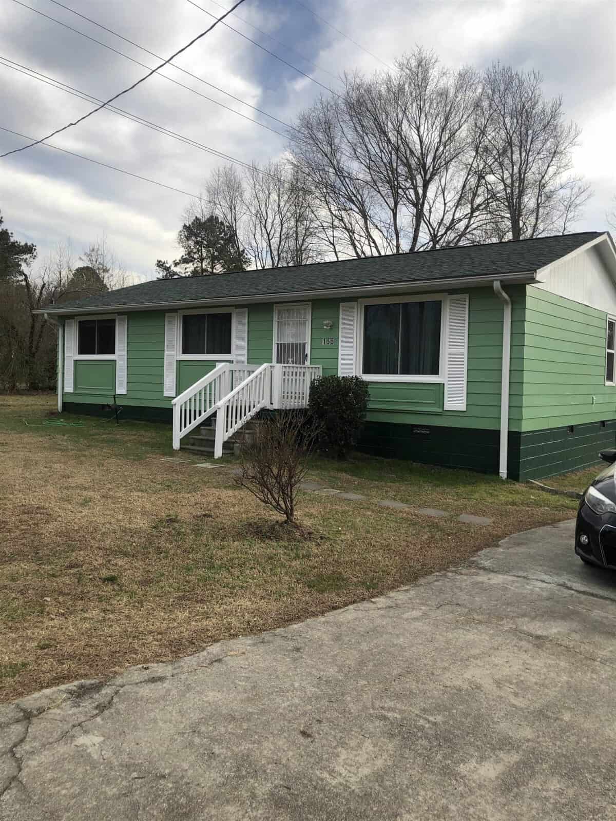 Single-story, green-painted house with white trim and a dark gray roof at 155 Kittrell Vance Avenue. The home has a front porch with white railings, steps leading to a white front door, and large windows. It is set against a grassy yard with bare trees and a driveway in the foreground in Kittrell.
