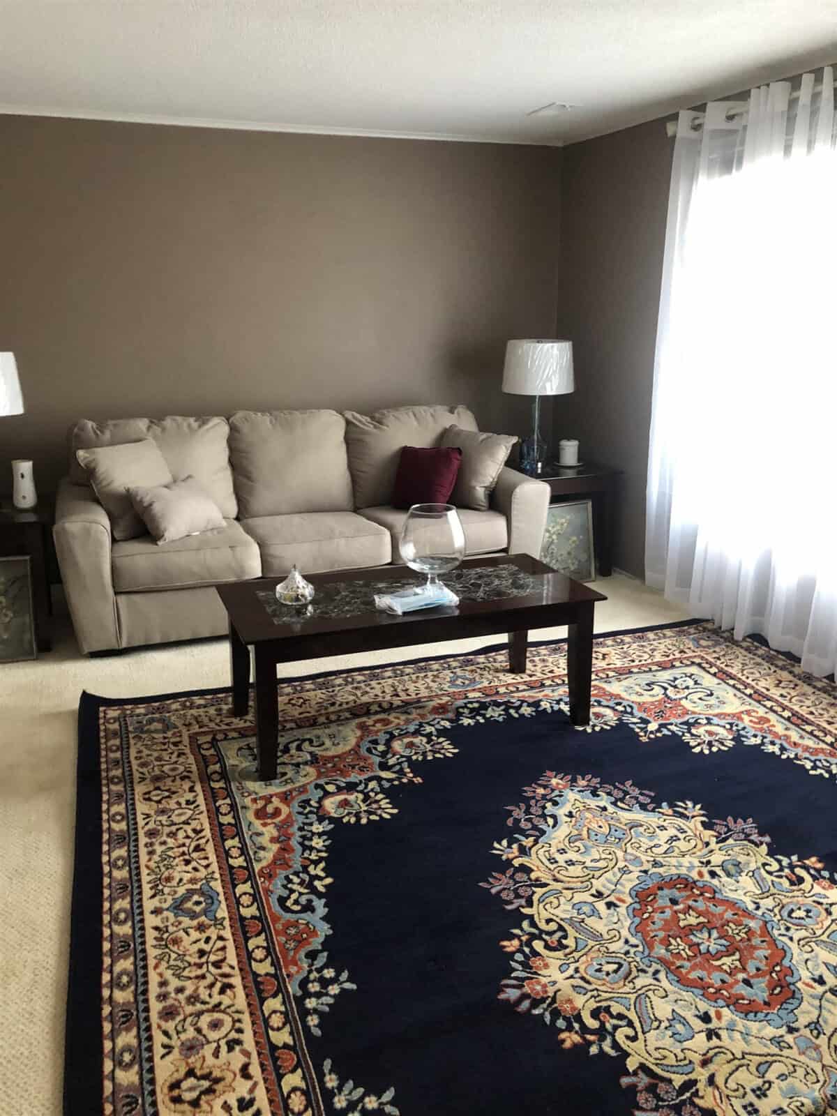 A living room with brown walls features a beige couch adorned with beige and burgundy cushions. Two side tables with lamps flank the couch. A dark wood coffee table is centered on an ornate blue and beige rug. White curtains allow natural light to stream in from the window.