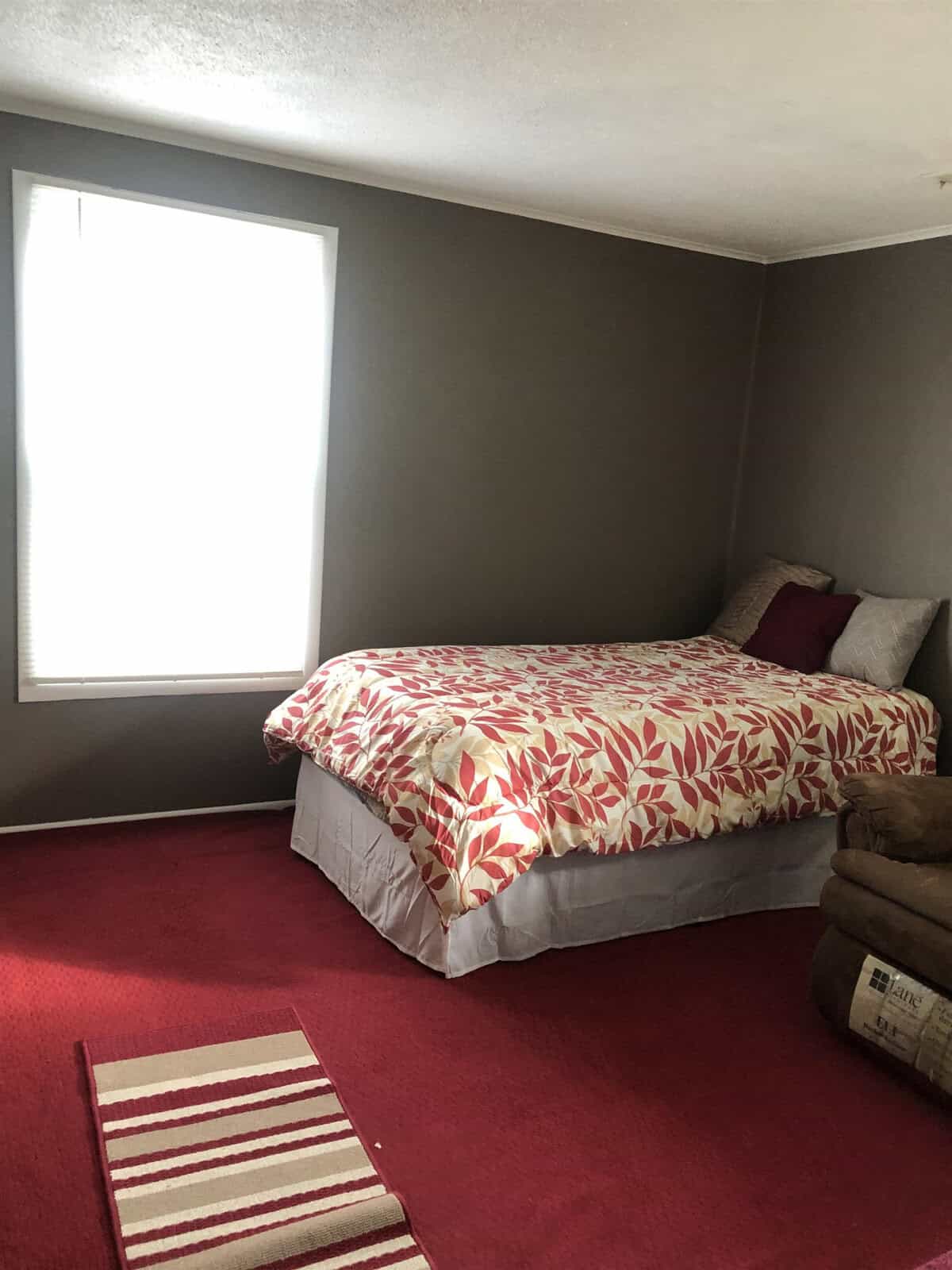 A small bedroom with a single bed pushed against the wall under a window. The bed has a red and white leaf-patterned comforter and several pillows. The floor is covered with a red carpet, and there is a red and white striped rug in front of the bed.