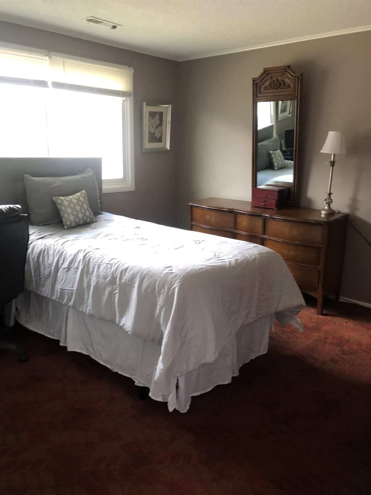 A cozy bedroom with a single bed featuring a white comforter and pillows, a cushioned headboard, and a wooden dresser with a mirror. A table lamp sits on one side of the dresser. A window with blinds provides natural light to the room. A framed picture hangs on the wall.