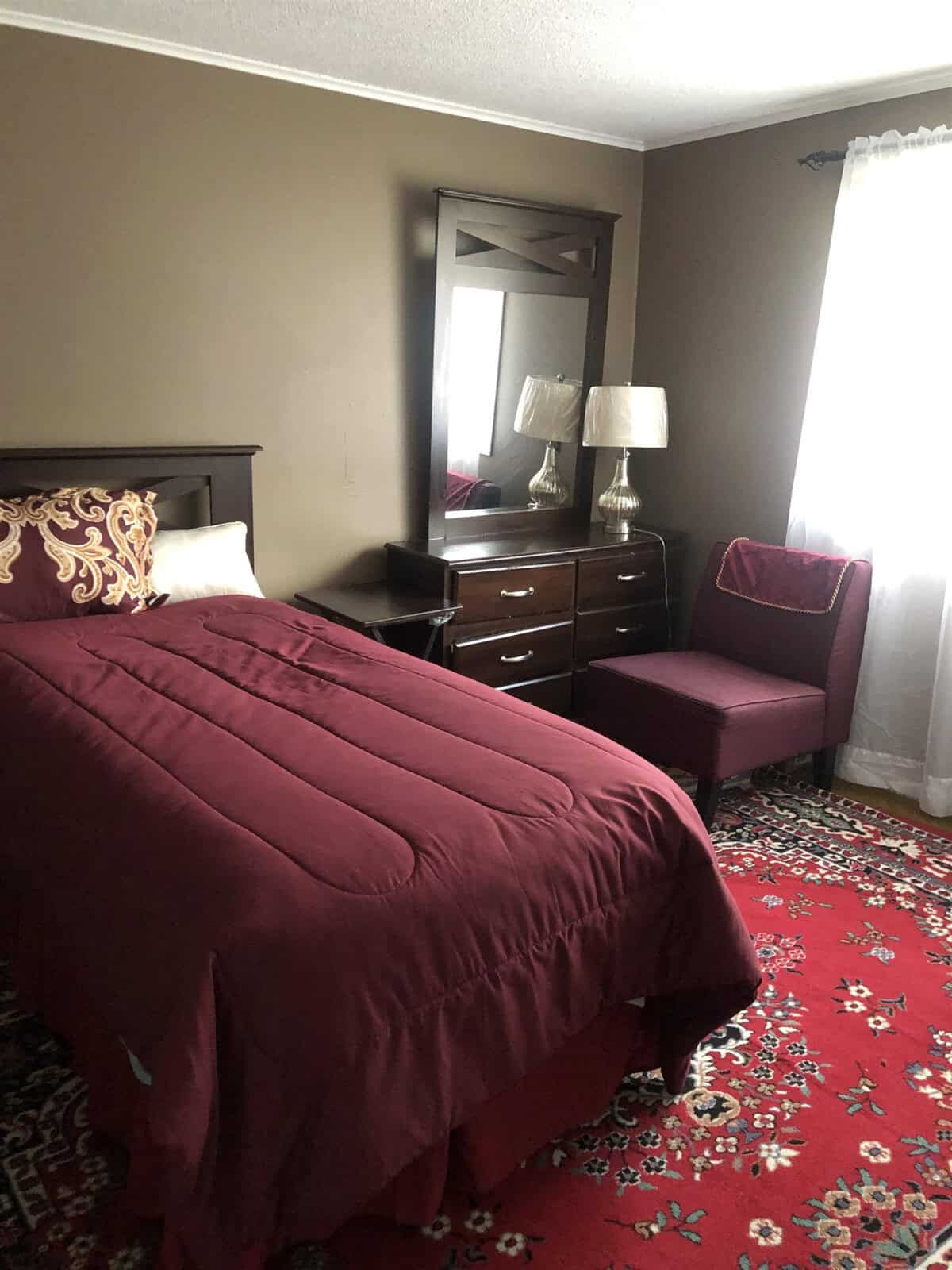 A cozy bedroom with a single bed adorned with a burgundy comforter and decorative pillow. The room features a dresser with a mirror, two table lamps, a chest of drawers, a burgundy upholstered chair, and a window with sheer white curtains. The floor has a patterned red carpet.