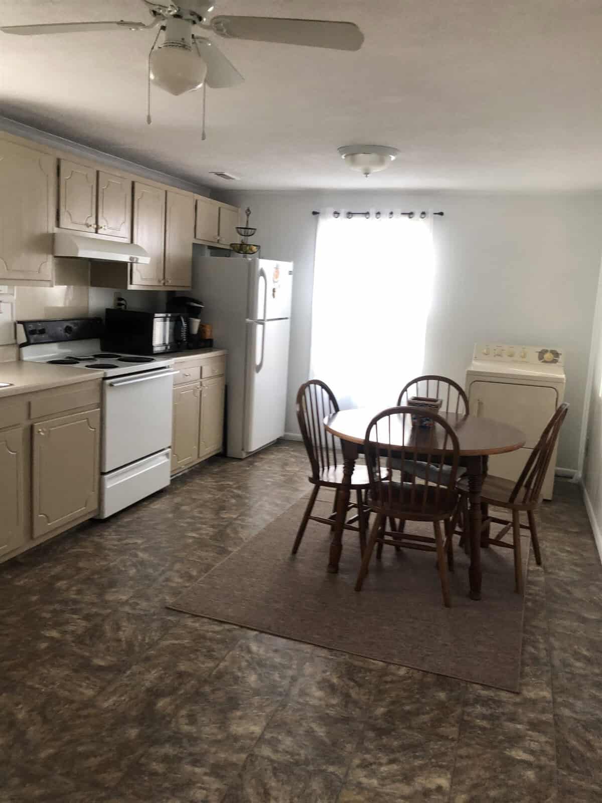 A kitchen with beige cabinets, a white fridge, stove, and microwave on the left side. A round wooden dining table with four matching chairs sits on a rug in the center. A white washer stands against the right wall near a window with curtains.