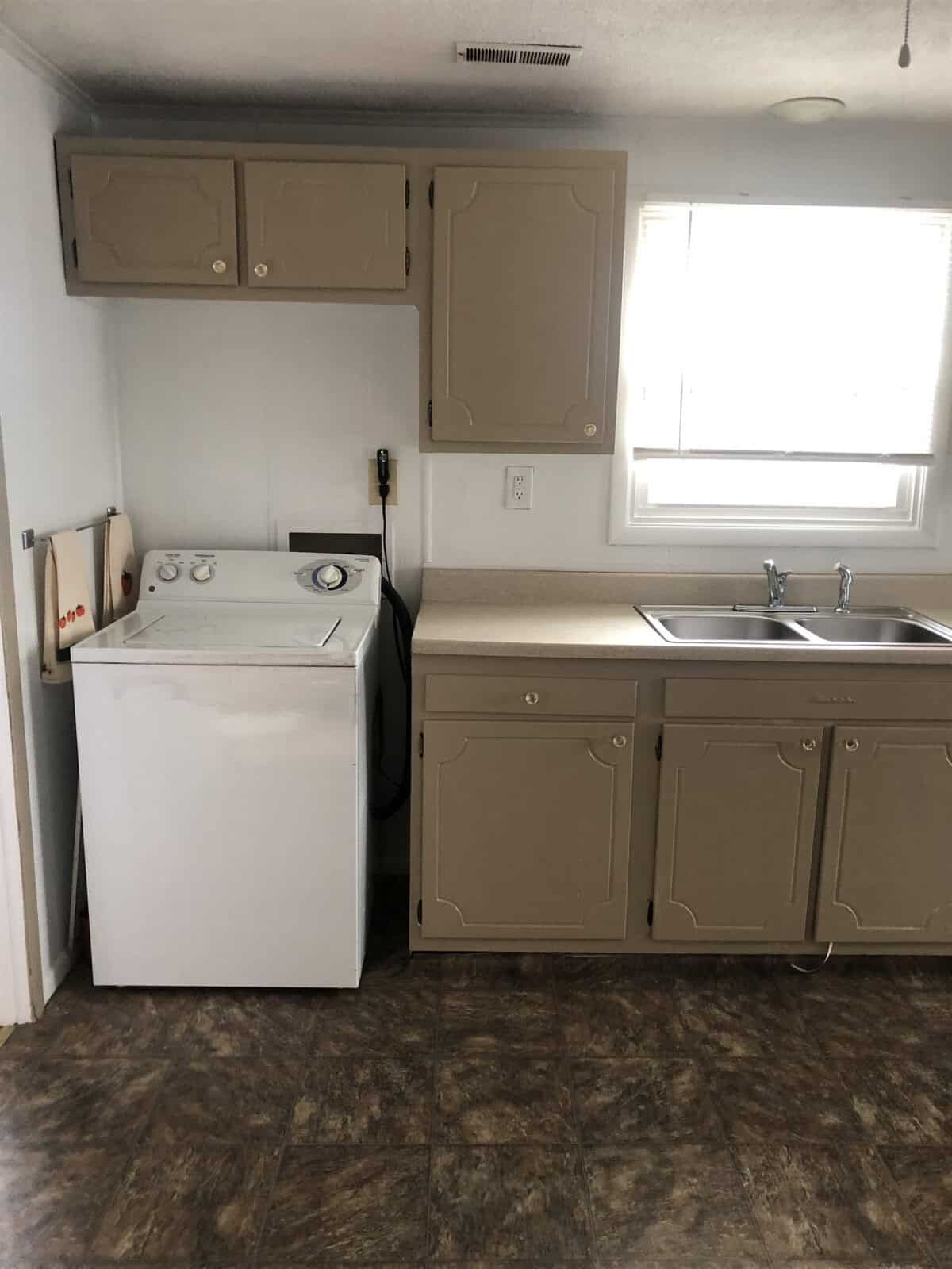 A small laundry area in a kitchen with beige cabinets, a white washing machine, and a double sink below a window with closed blinds. A handheld shower head is hanging on the wall next to the washing machine. The floor is covered in patterned linoleum.