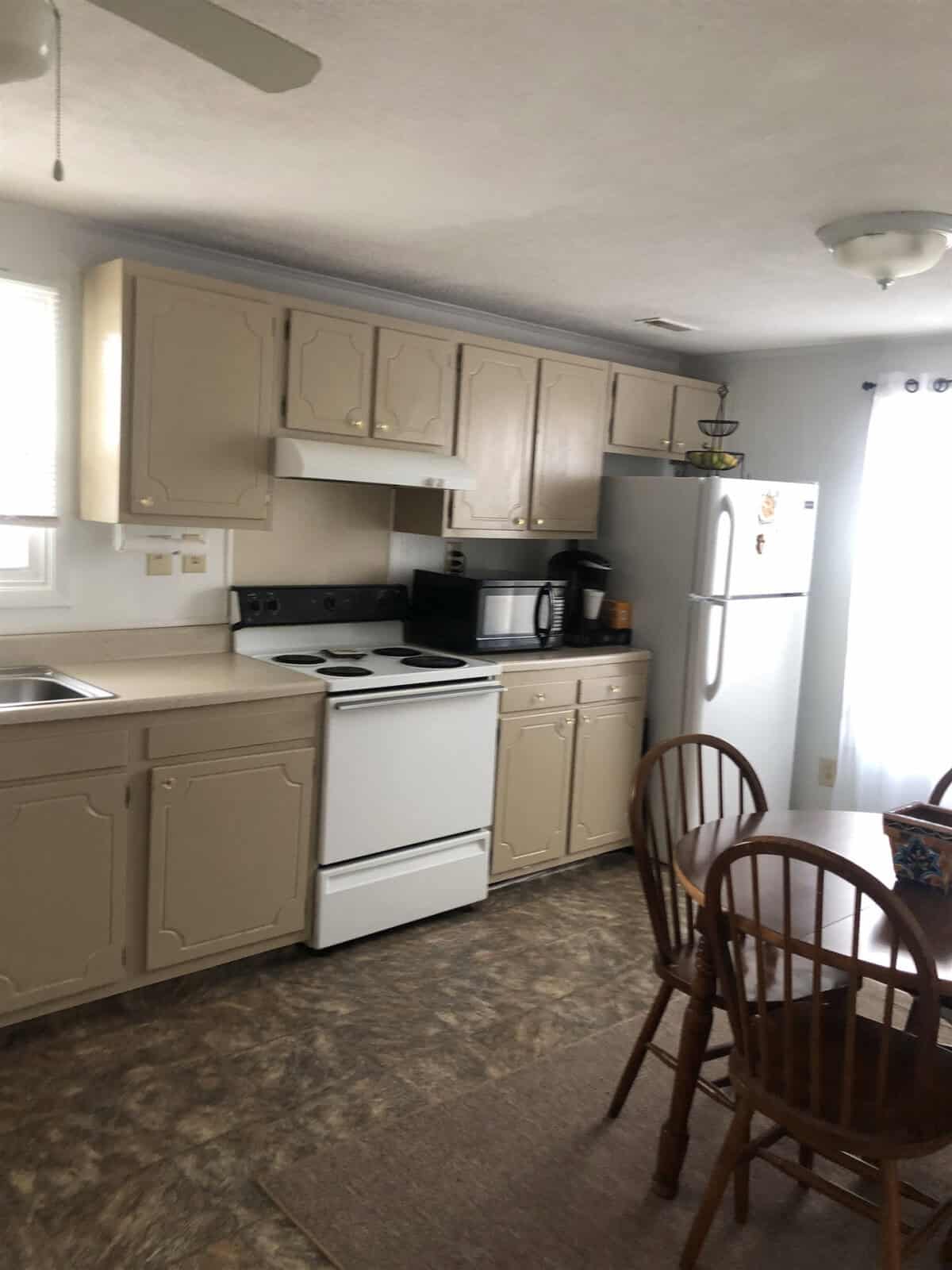 A kitchen with beige cabinets, a white stove, and a white refrigerator. There is a microwave on the counter and a sink under a window with light coming in. Adjacent to the kitchen area, there is a small dining table with two wooden chairs. The floor is vinyl.