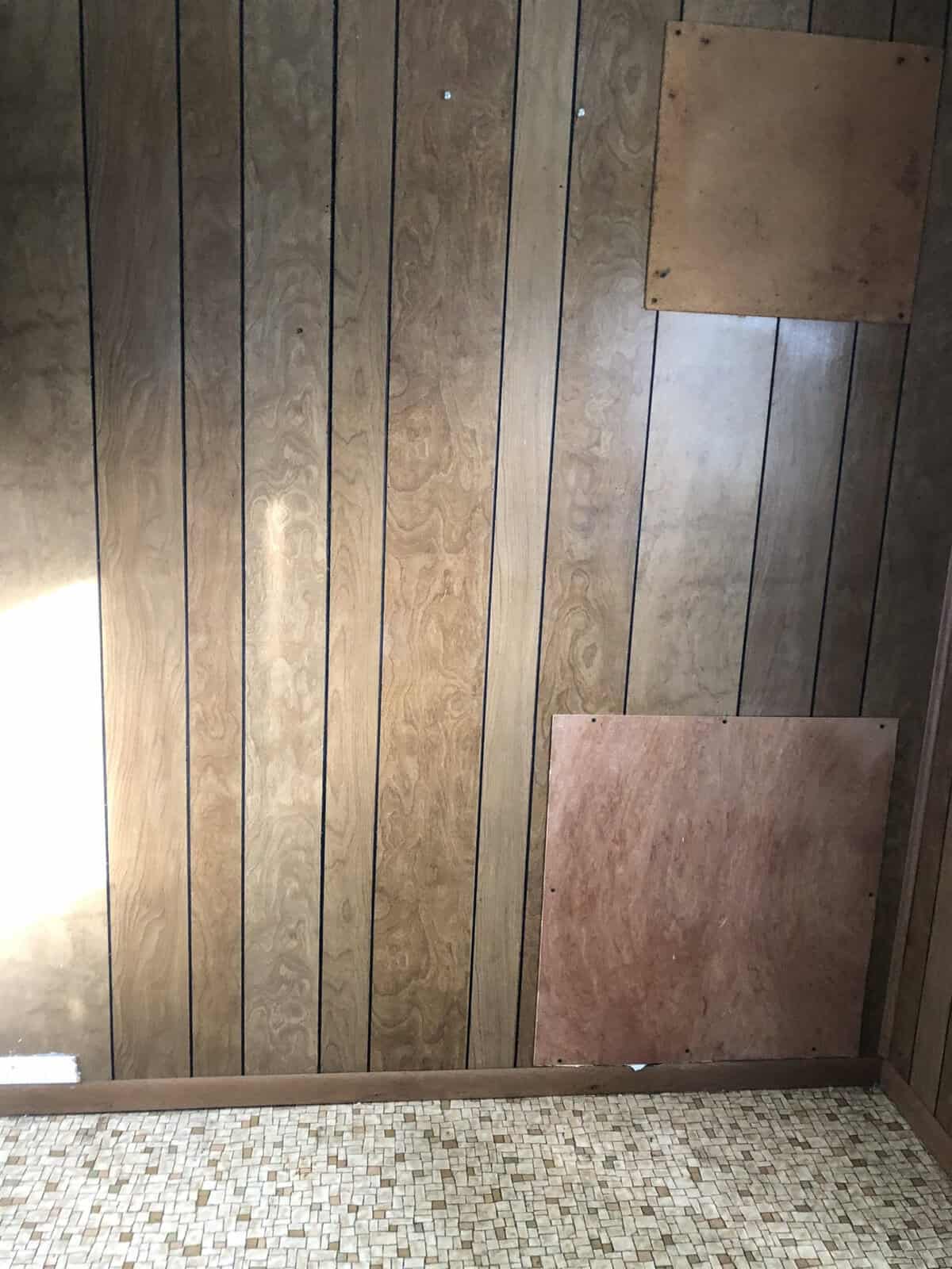 A room with wood-paneled walls and patterned linoleum flooring. The wall has two square patches of different wood, indicating repairs or covering. Sunlight partially illuminates the wall from the left side.