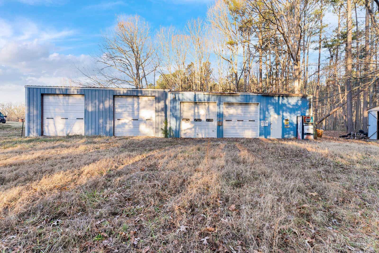 A blue metal storage building with four garage doors, situated on a grassy field with trees in the background and a clear sky above, located at 1150 Gun Club Road, Henderson NC.