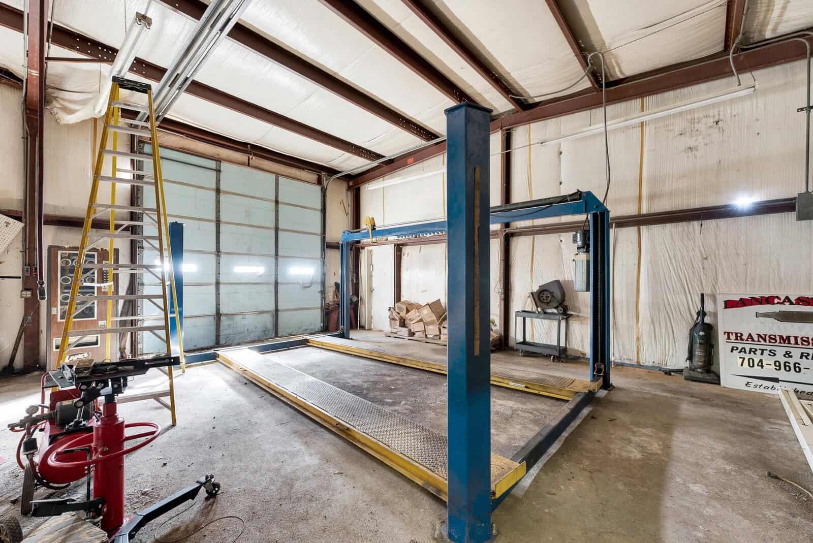 A spacious garage workshop with an automotive lift, a transmission jack, a ladder, and various other tools and equipment scattered around the area.
