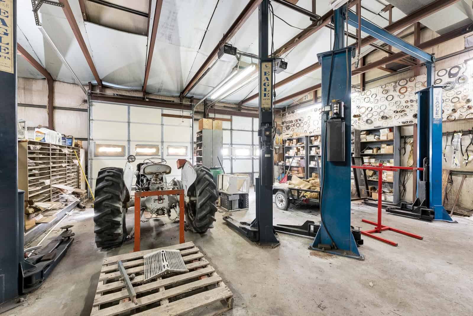 A spacious workshop with two vehicle lifts, a tractor being serviced, various tires, tools, and equipment, and a neat stack of wooden planks towards the back.