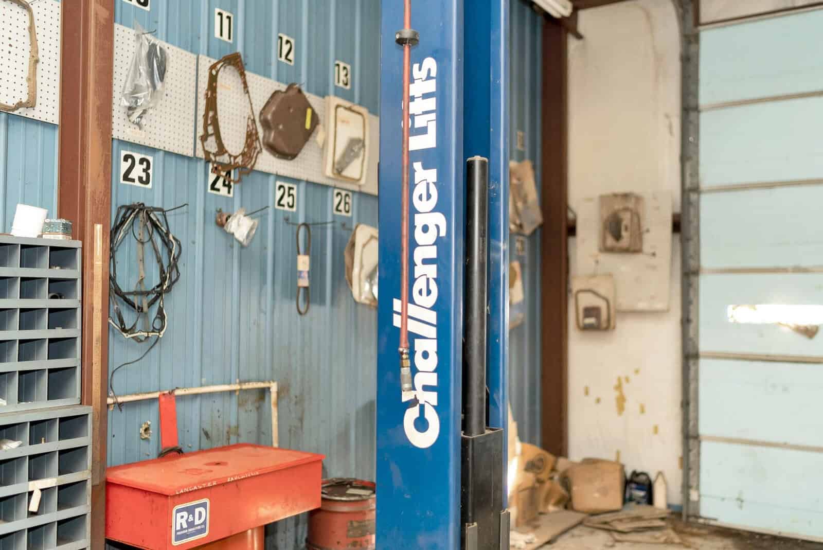 A vehicle lift labeled "challenger lifts" stands on the right side of a cluttered automotive repair garage, with various tools and equipment scattered around the space.