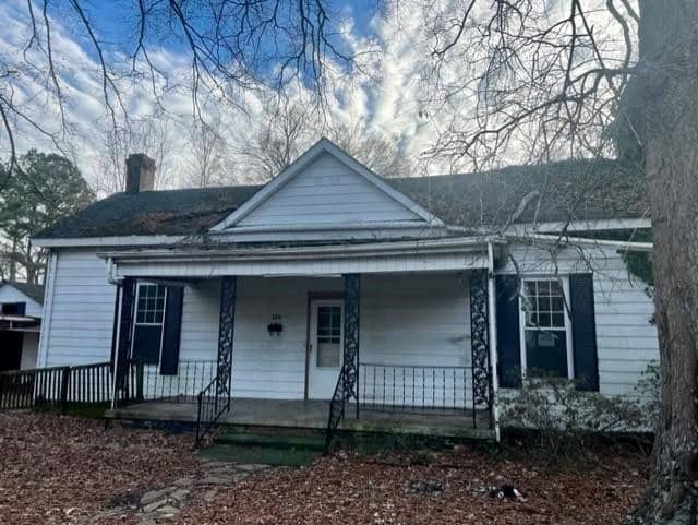 A weathered, white single-story house with dark shutters, a central chimney, and visible signs of neglect, including a sagging gutter and overgrown yard.