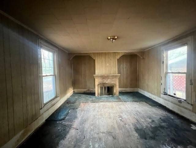 An old, empty room with worn-out wooden paneling, a fireplace, and two large windows letting in natural light. the floor appears damaged and there is a discolored carpet remnant near the hearth.