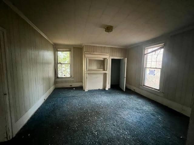An empty room with white paneled walls, a dark carpet, and a built-in cabinet next to a fireplace, illuminated by natural light from windows.