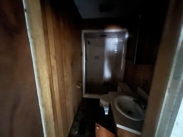 A small, dimly lit bathroom with wooden paneling featuring a white toilet and a shower stall with a glass door.