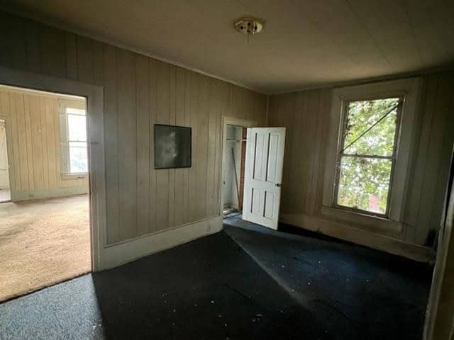 A dimly lit empty room with paneled walls, a single ceiling light fixture, dark carpeting, and a detached white door leaning against a wall next to a closed window.