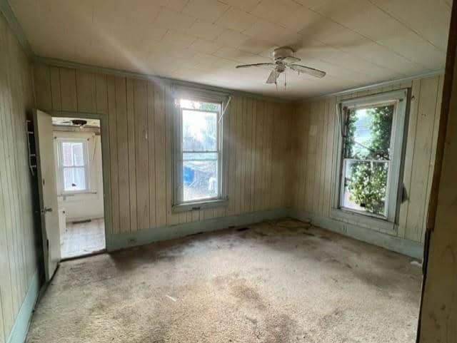 An empty room with worn carpet, paneled walls, and a ceiling fan, featuring a window that overlooks the outdoors and an open doorway leading to another room.