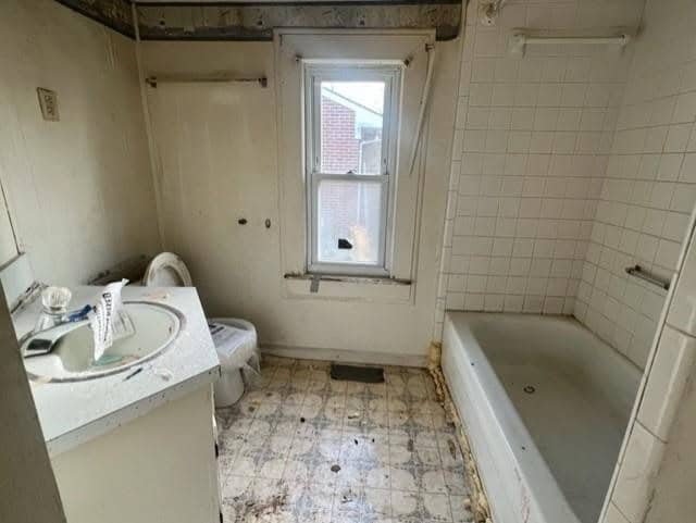 A dated and unclean bathroom featuring a dirty tiled floor, a sink with scattered personal items, a tub, and a window with a partially opened blind.
