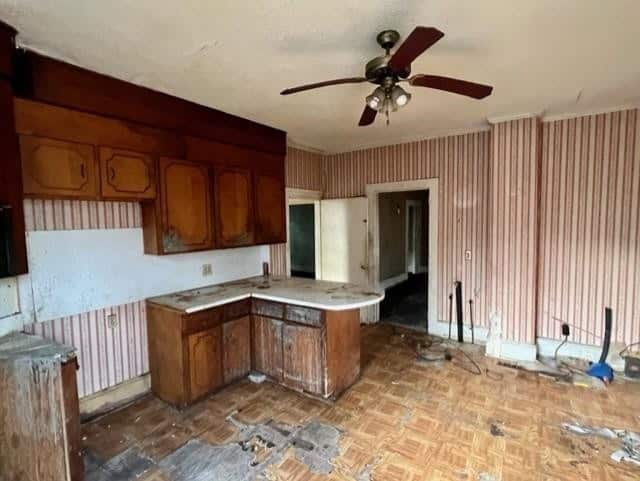 An old, dilapidated kitchen with damaged parquet flooring, a ceiling fan, and striped wallpaper.