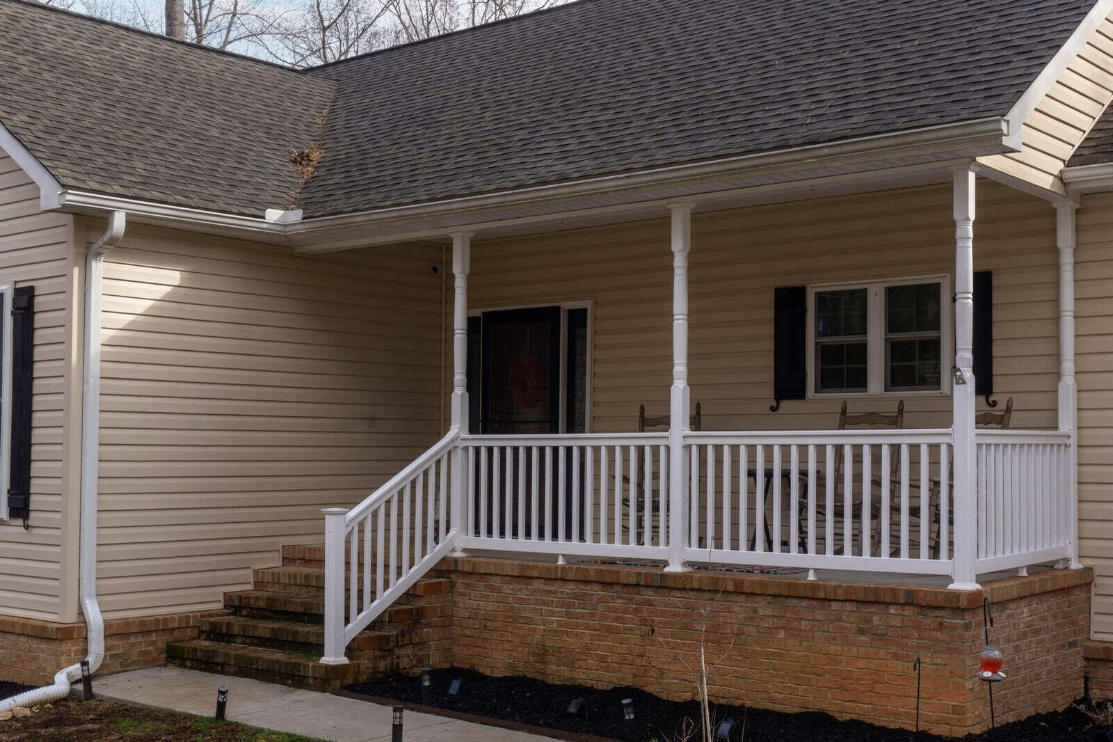 A residential home featuring a covered front porch with white railings, accessed by a set of brick and wooden stairs, and a beige siding exterior.