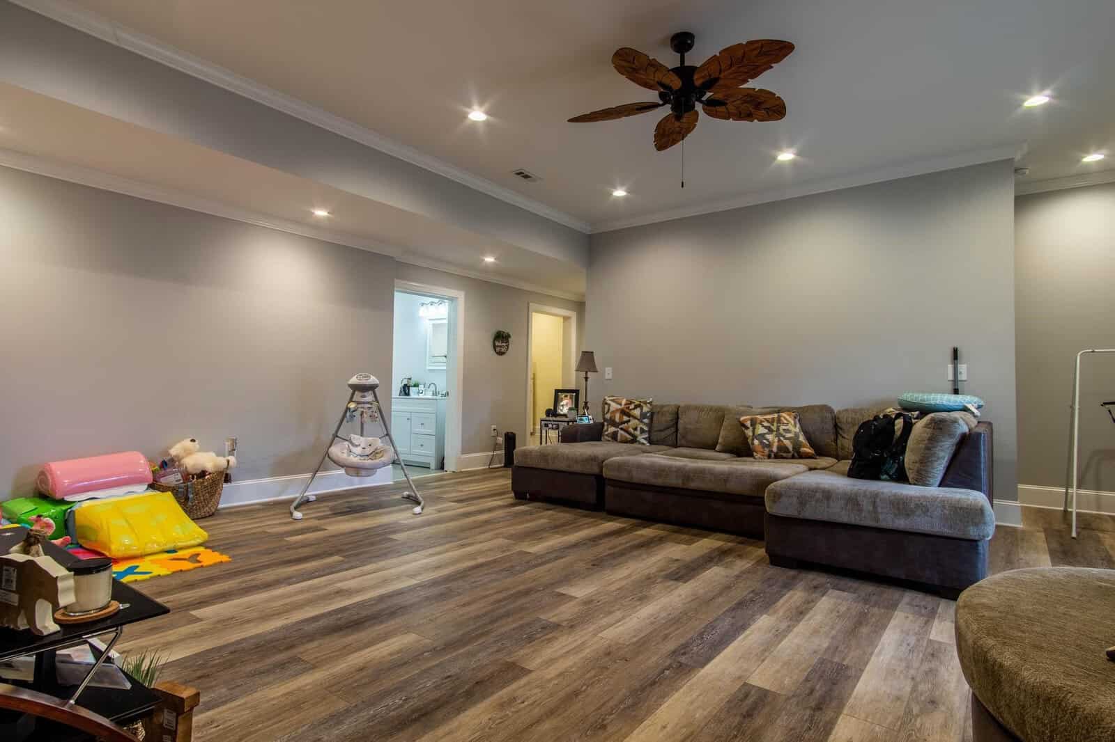A spacious living room with hardwood flooring, recessed lighting, a sectional sofa, and various baby items including a swing and play mat.
