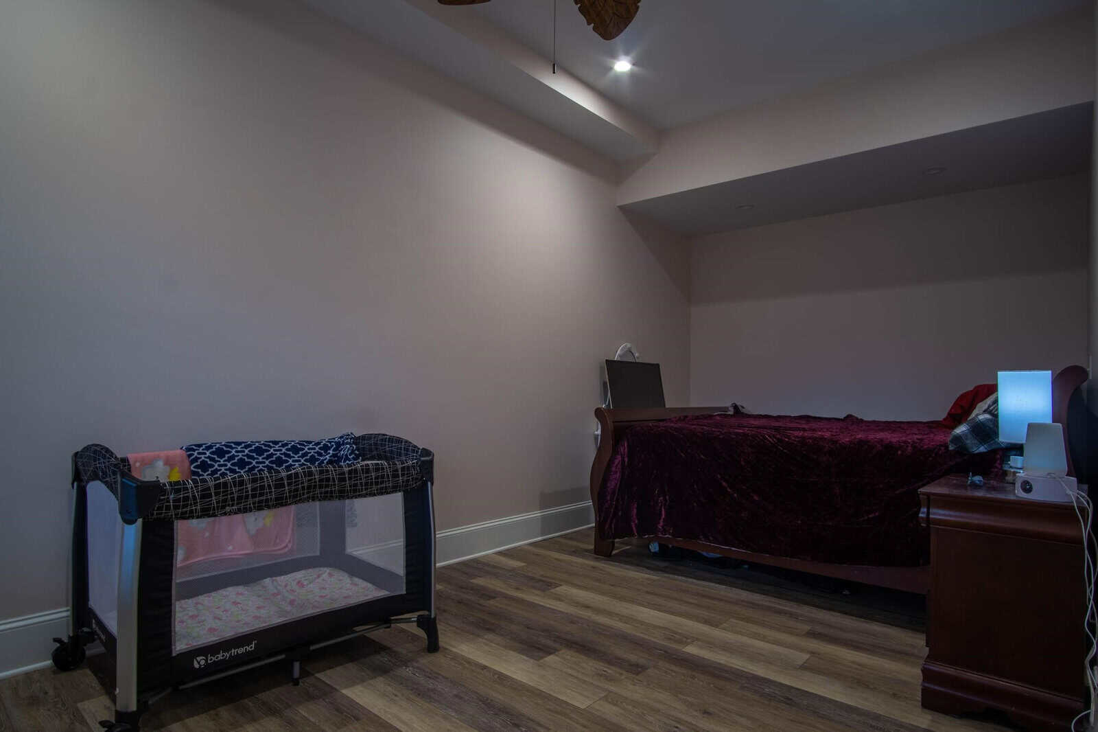 A bedroom with a wooden floor featuring a twin bed with burgundy bedding on the right, a portable baby crib on the left, and a desk with a computer against the far wall.