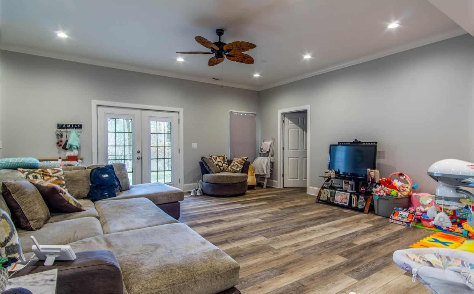 A spacious living room with modern decor, featuring a large l-shaped sofa, a ceiling fan, recessed lighting, and a hardwood floor. the room includes a television, a variety of children's toys scattered on the floor, and a separate seating area with patterned armchairs near the window.