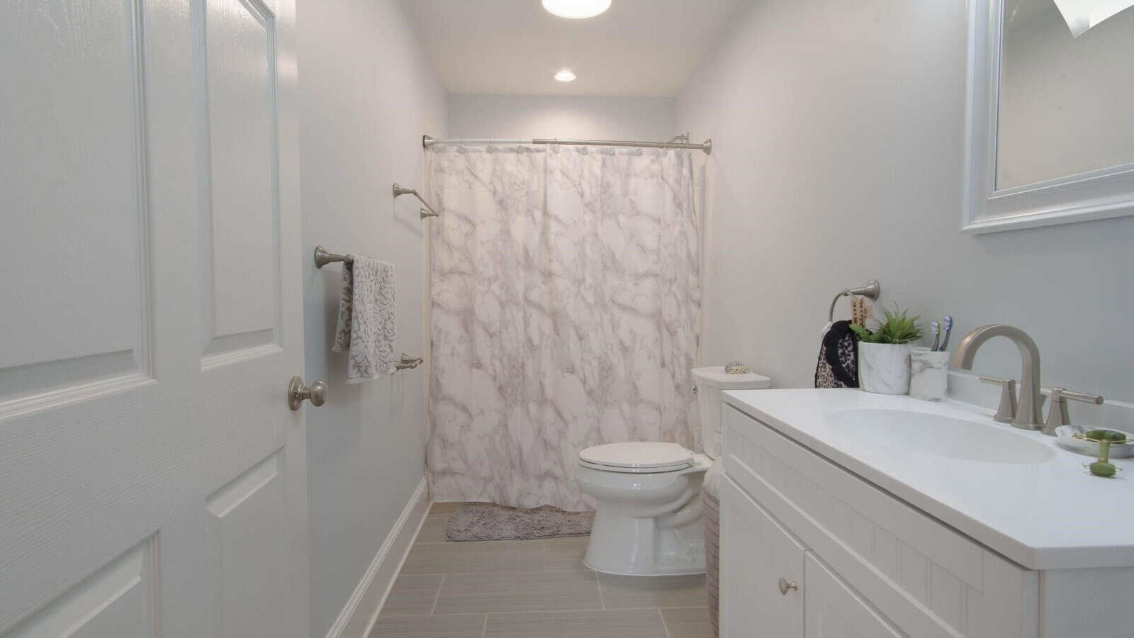 A clean and modern bathroom with a white interior, featuring a toilet, vanity sink, shower with a marble-patterned curtain, and a gray bath mat.