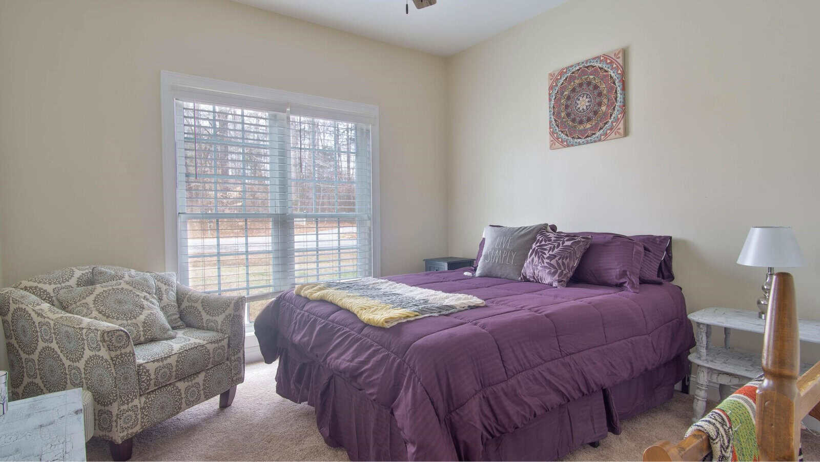 A neatly arranged bedroom with a purple bedspread, patterned armchair, a lamp on a bedside table, and a decorative wall hanging above the bed.