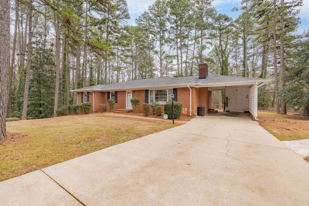Single-story brick house with a carport nestled among tall pine trees.