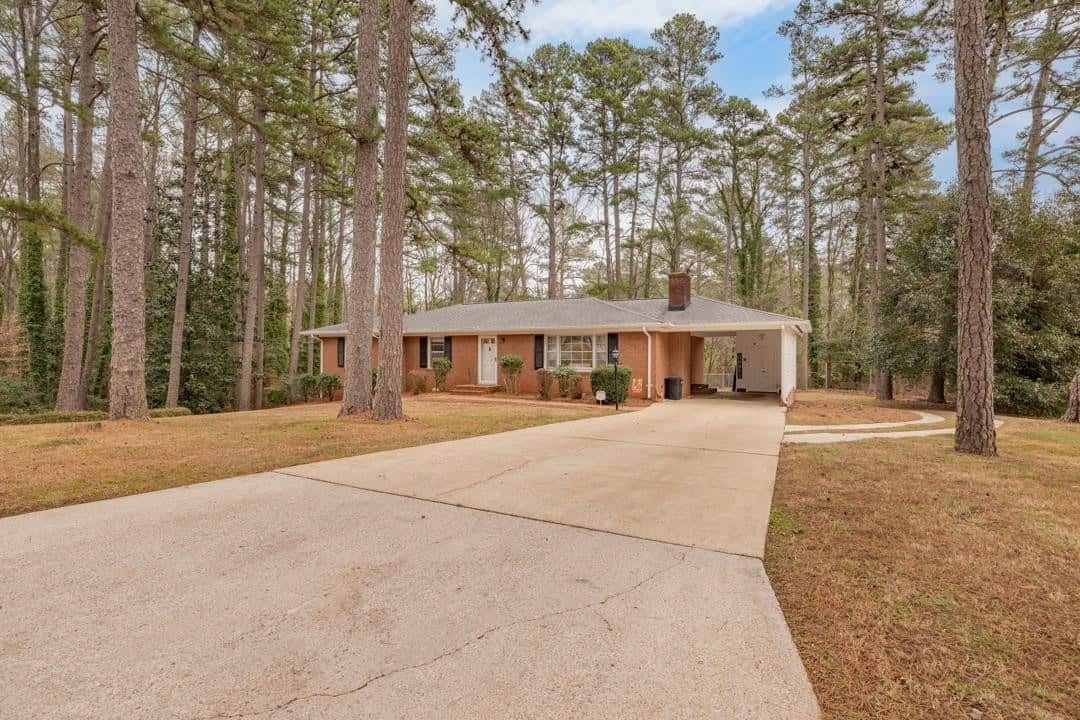A single-story brick house with a white trim, surrounded by tall pine trees, featuring a large concrete driveway that leads to an attached carport.