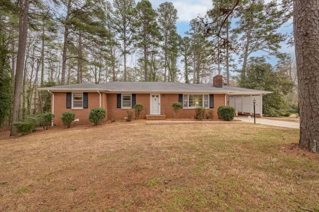 One-story brick house with white trim, a chimney, and an attached garage situated in a yard with pine trees.