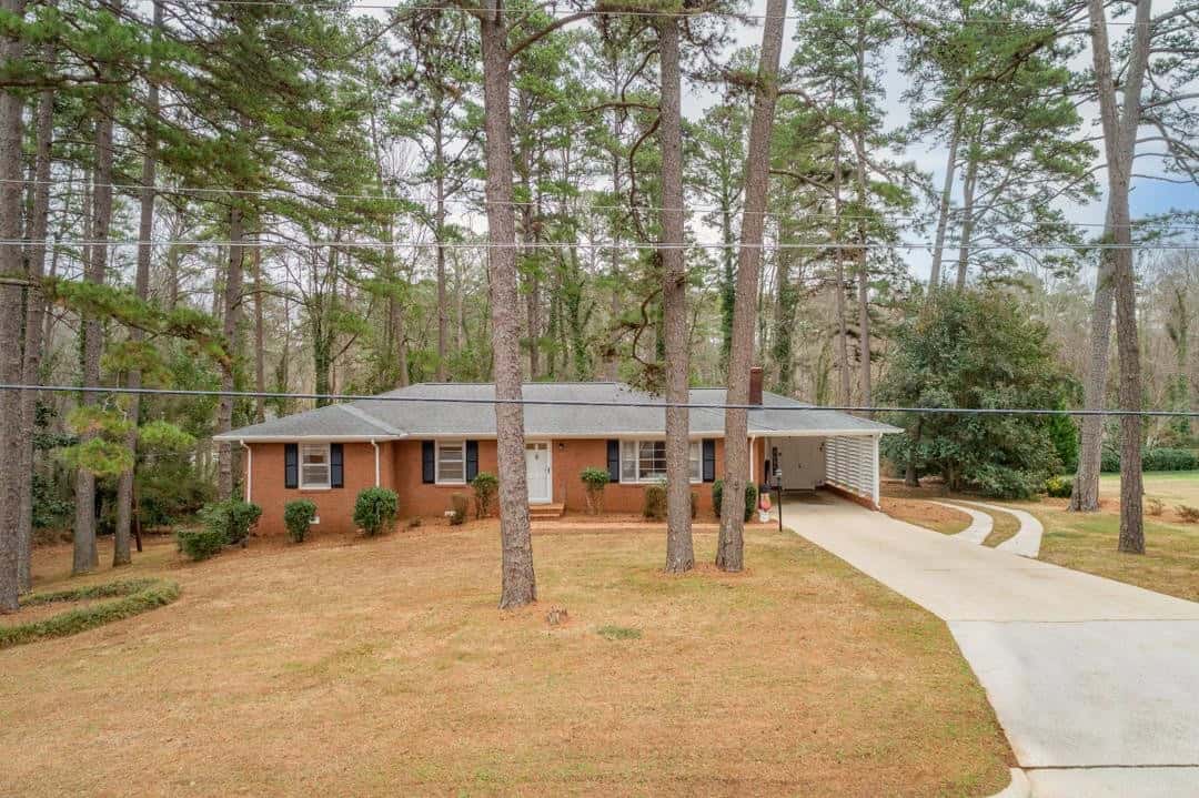 A single-story brick house set amidst tall pine trees with a concrete driveway leading to an attached carport.
