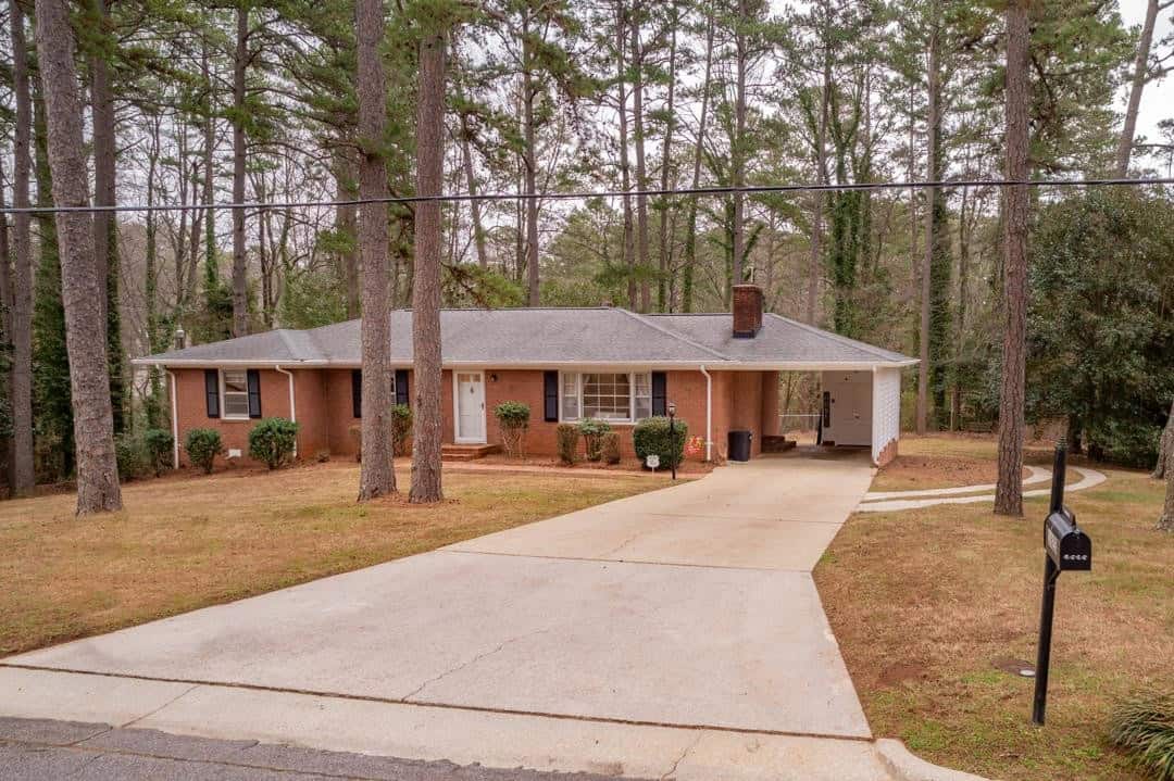A single-story brick house with a gabled roof, flanked by tall pine trees, featuring a driveway leading to an attached carport on the right, under a clear sky with thin cloud cover. a black mailbox stands by the curb in the foreground.
