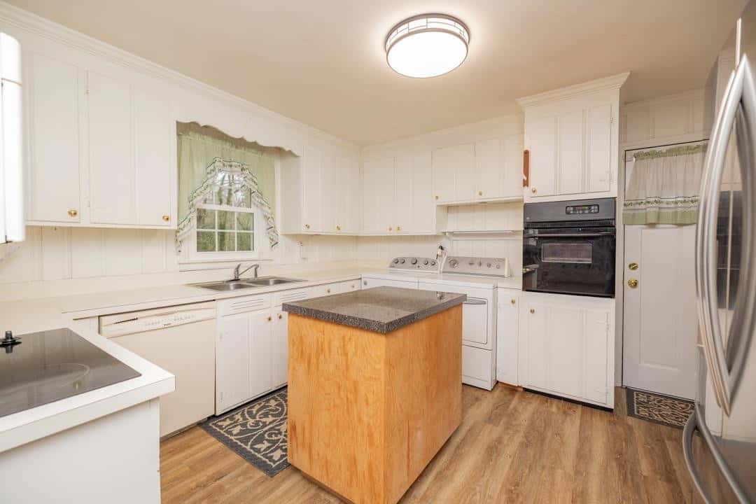 A spacious kitchen with white cabinetry, laminate countertops, and hardwood flooring, featuring a central island, white appliances, and a window with patterned curtains.