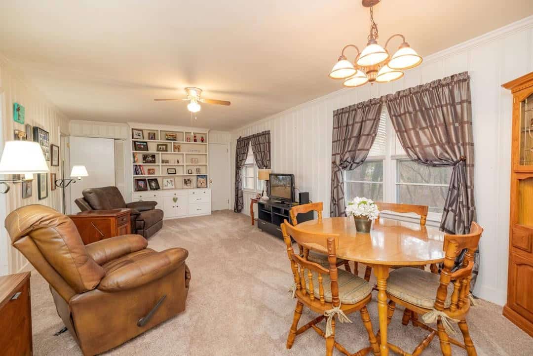 A spacious living room furnished with a brown recliner chair, a wooden dining set, and a television set on a stand, featuring carpeted flooring, white walls with decorative items, and curtained windows allowing natural light.