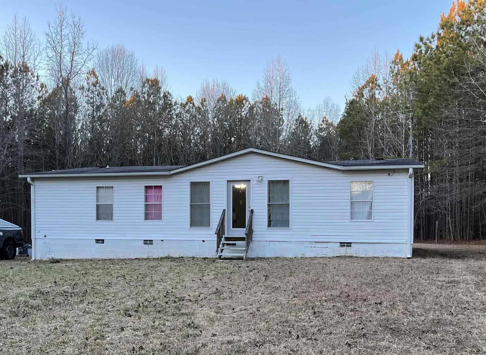 A single-story white mobile home with a black roof, featuring a central door flanked by two windows, one with a bright pink curtain, set against a backdrop of trees with a vehicle parked to the left.