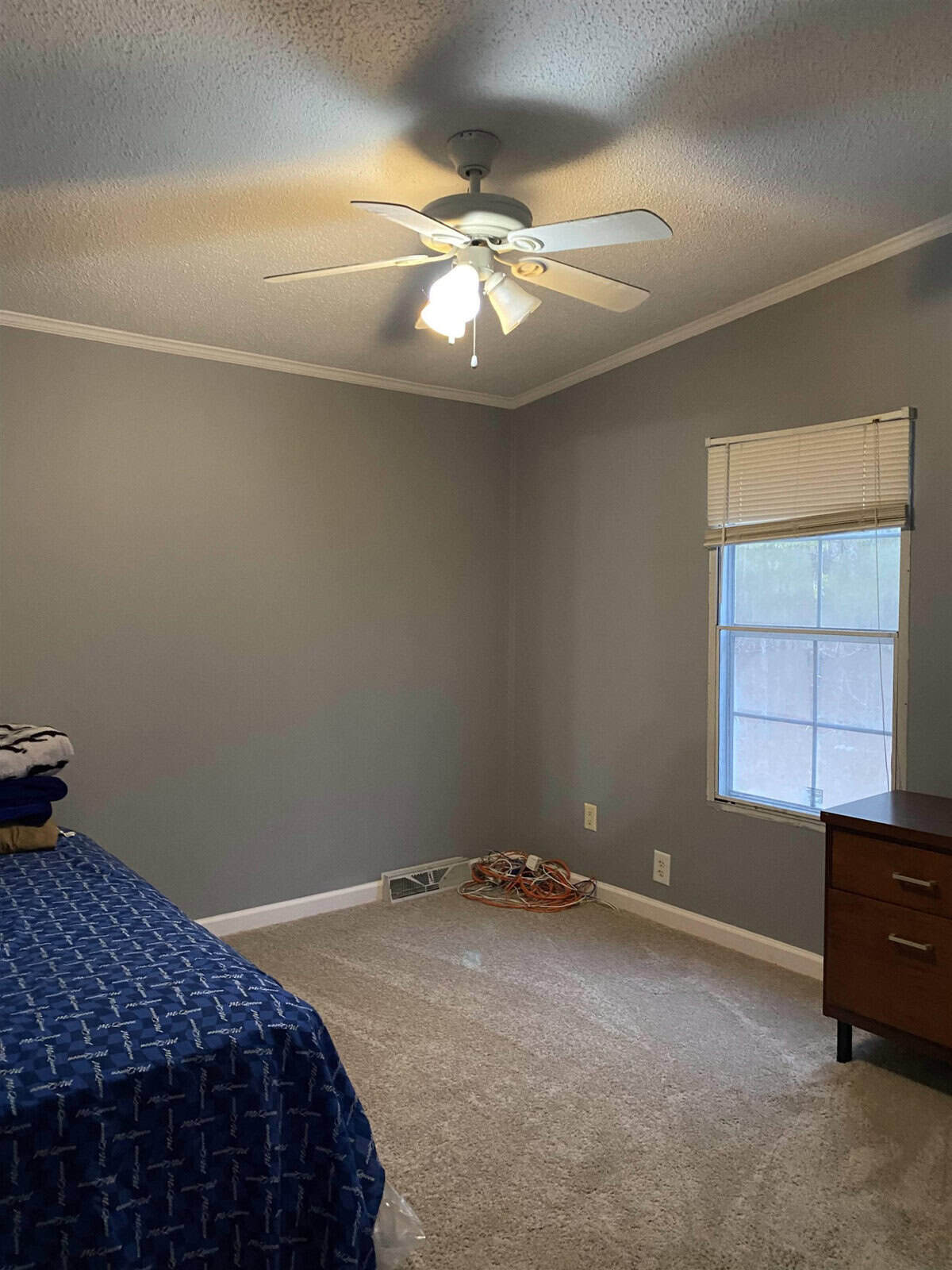 A sparsely furnished room with gray walls featuring a bed with a blue cover, a ceiling fan with lights, a wooden nightstand, and a window with blinds. a pile of orange extension cords lies on the floor.