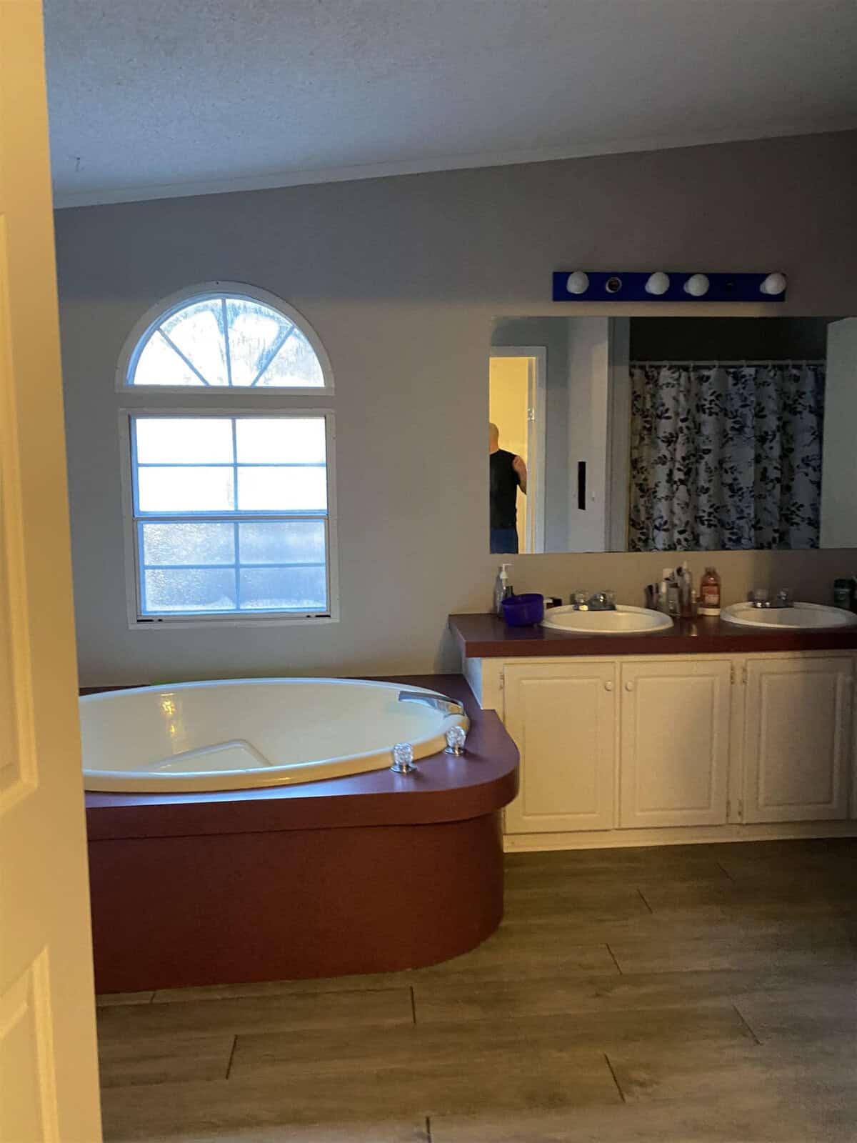 A residential bathroom with a corner bathtub beneath a window, adjacent to a vanity with a sink and a mirror, with a frosted glass window providing natural light. there is a person partially visible in the adjoining room.