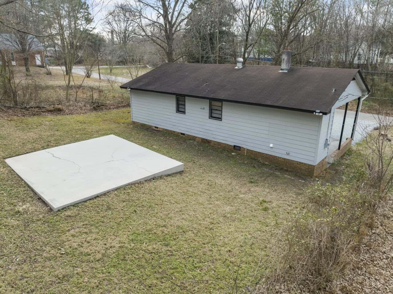 A single-story house with a white siding exterior, a dark shingled roof with two chimneys, and surrounded by a grassy yard with a large, concrete slab in the foreground.