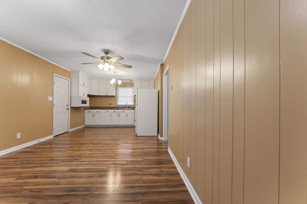 An empty kitchen with wooden flooring, white appliances, and beige walls, featuring a ceiling fan with lights.
