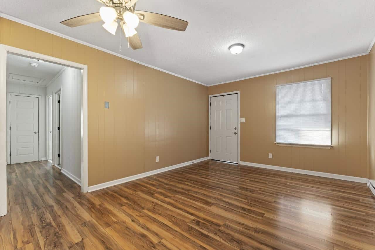 A well-lit empty room with wooden flooring, beige walls, a white door, and a ceiling fan.
