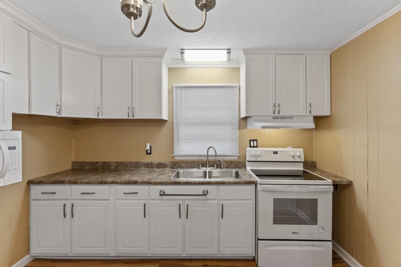 A compact kitchen with white cabinets, laminate countertops, and a suite of white appliances, including a stove and a dishwasher, with a window above the sink and beige walls.