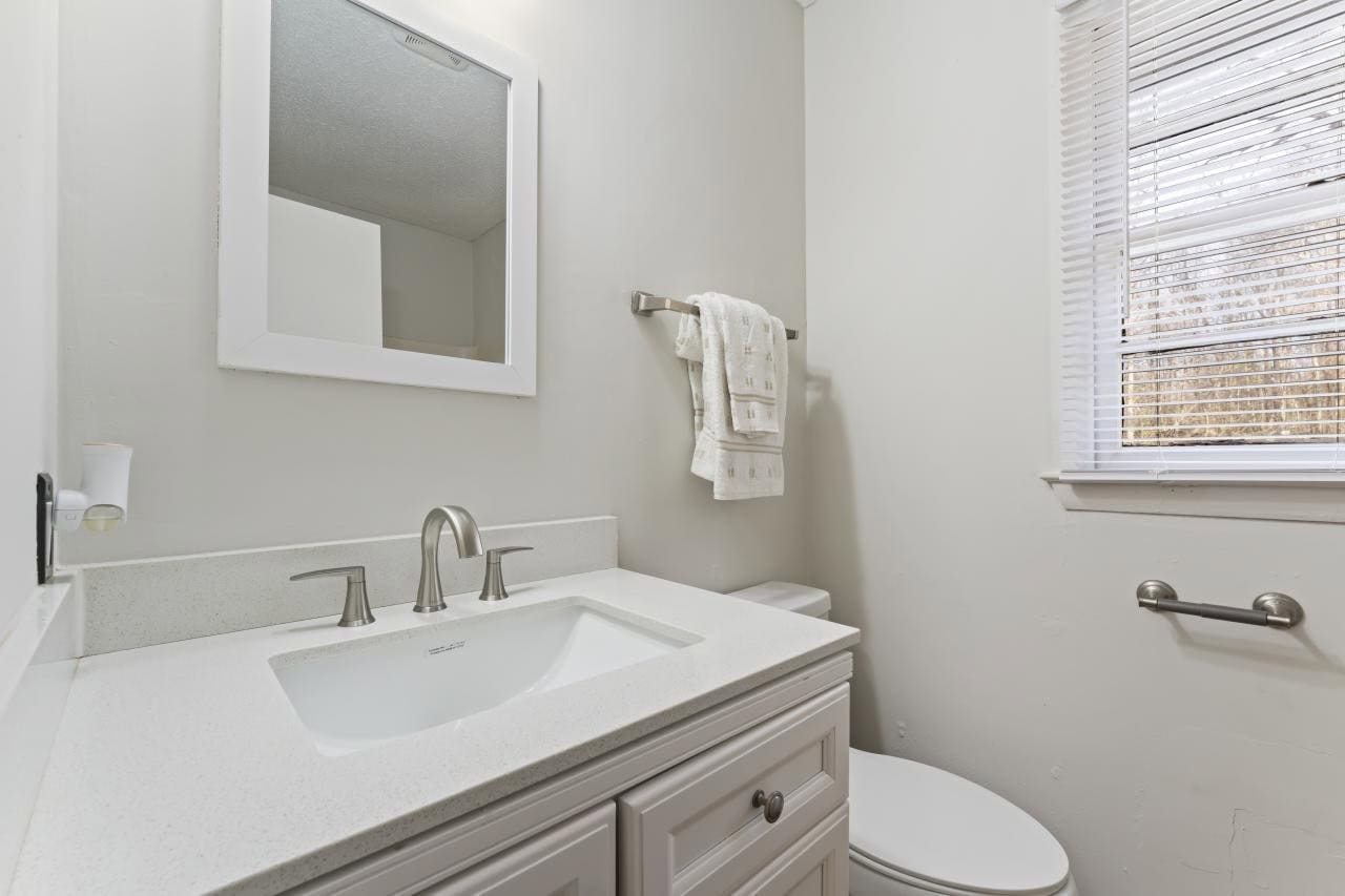A small, clean bathroom with a white vanity that includes a sink, a mirrored medicine cabinet above it, and a toilet to the right; a towel is hung on a rack, and there is a window with blinds allowing natural light in.