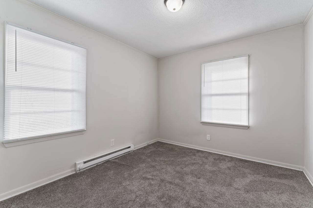 An empty room with white walls, two windows with blinds, and grey carpet flooring.
