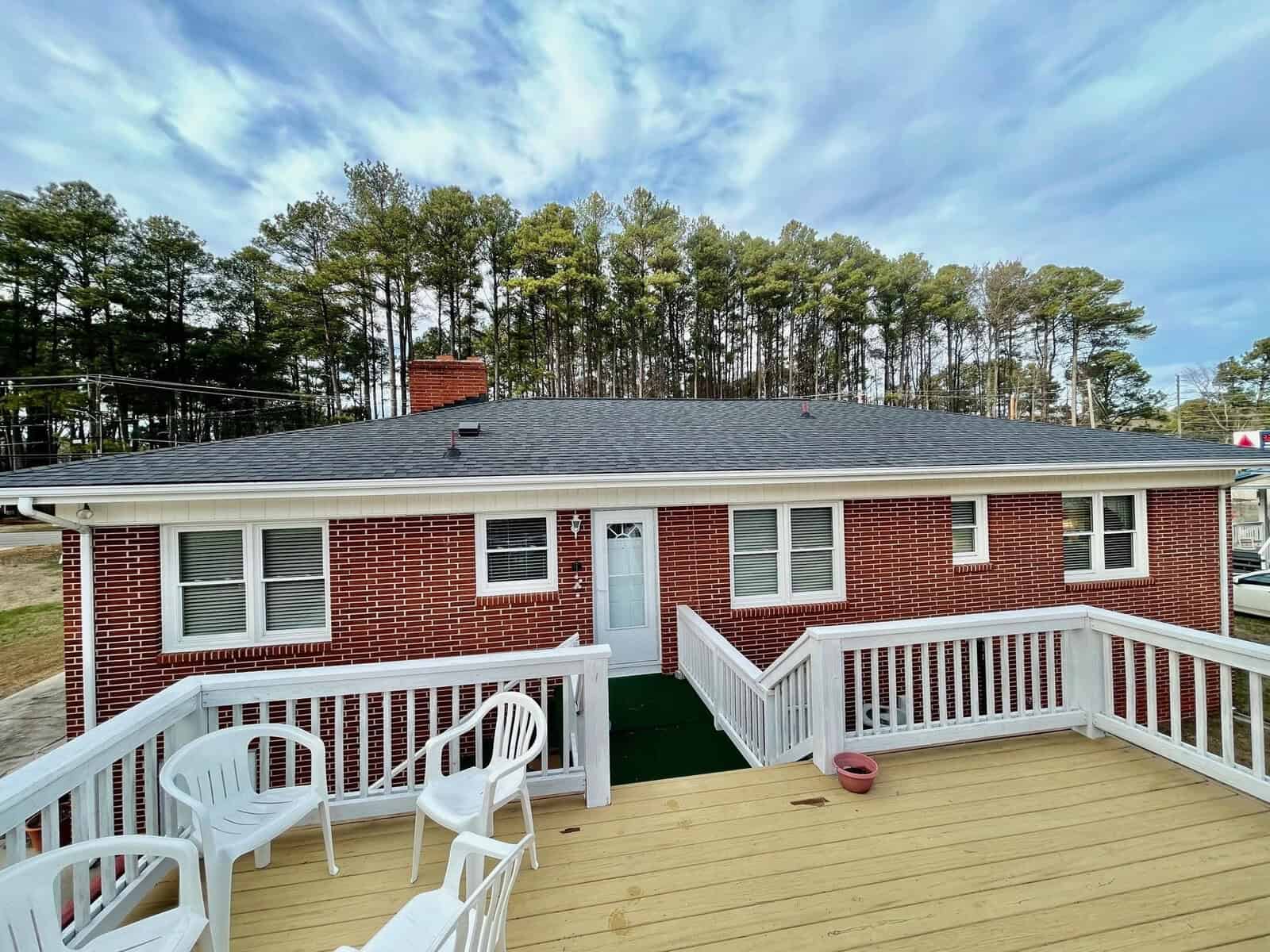 A single-story brick house with a dark shingled roof and a wooden deck, under a partly cloudy sky.