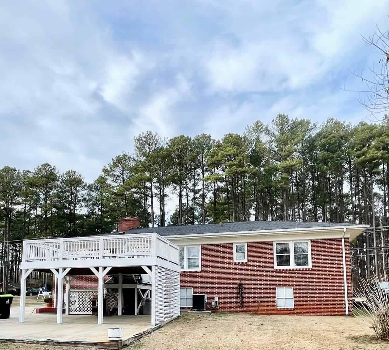 A red brick single-story house with a white elevated deck on the left side, set against a background of tall pine trees and a cloudy sky.