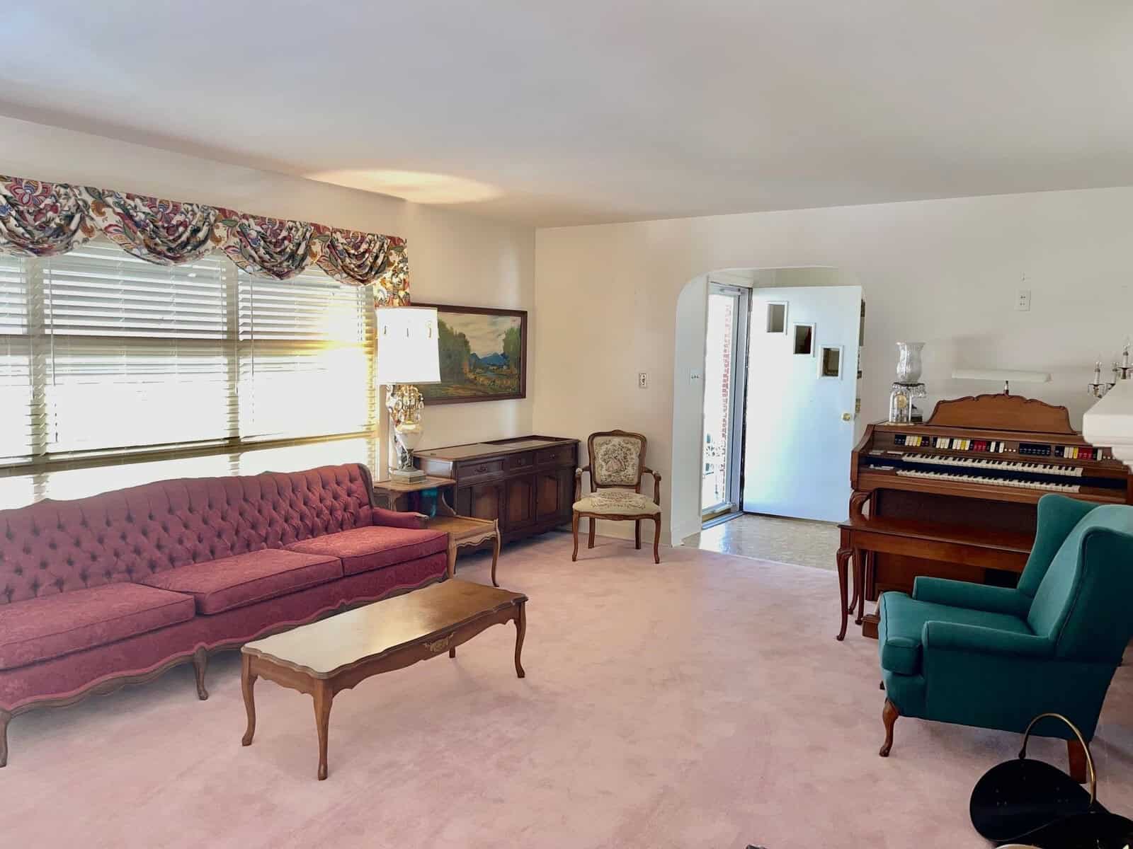 A well-lit, traditional living room featuring a long red sofa, a turquoise armchair, a wooden coffee table, and an upright piano by the wall. the room is carpeted in pink, and there is a painting above a small wooden table against the wall.