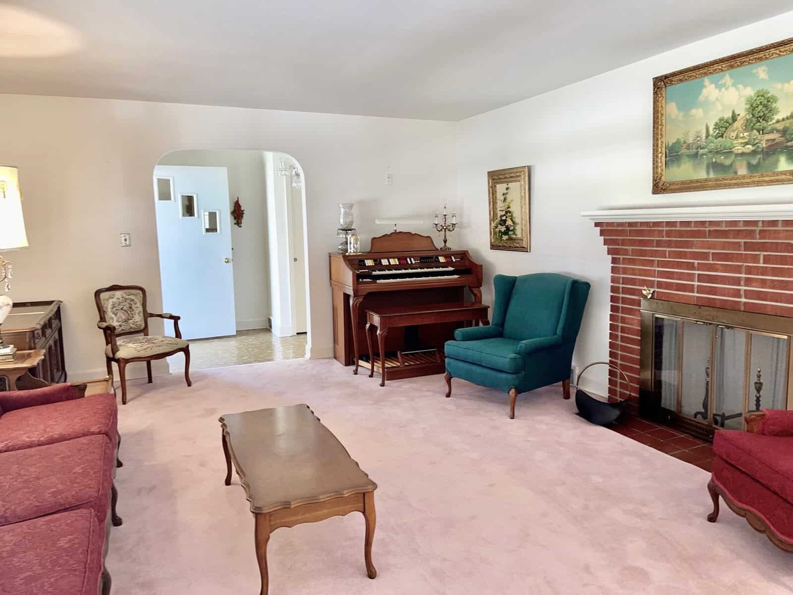 A traditional living room featuring a red brick fireplace, a blue armchair, a piano against the wall, and various other pieces of classic furniture, with framed artwork hanging above the fireplace and on the adjacent wall.