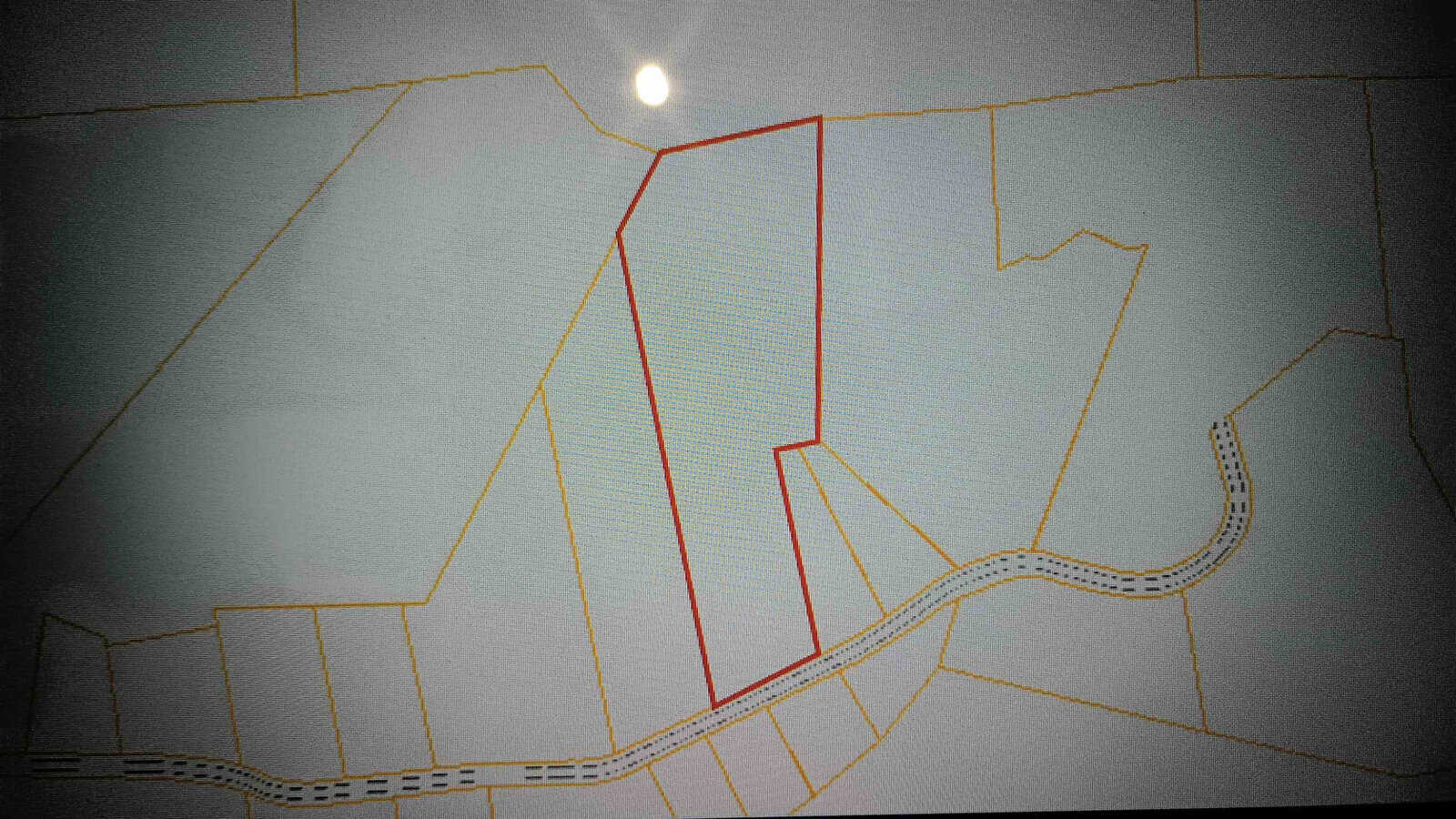 A grayscale topographic map with light grid lines is shown. A specific irregularly-shaped area is highlighted with a red outline. Several adjacent areas are marked with orange outlines, and a dotted road curves through the lower portion of the map.