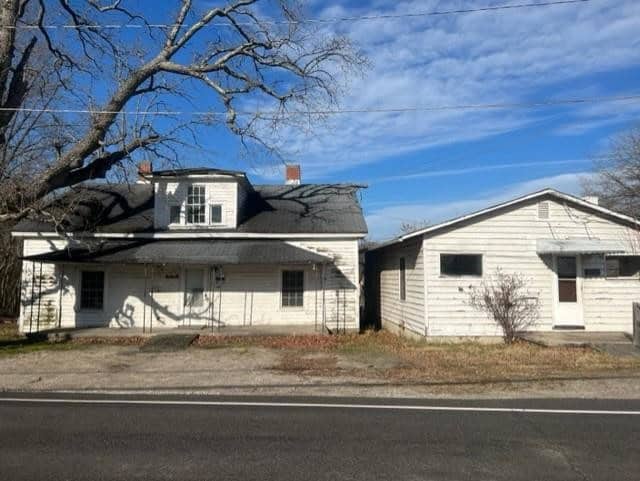 A deteriorating white house with a damaged roof and attached garage, situated at 1095 Old Norlina Road, Henderson NC, beside a road with leafless trees in the background.
